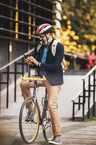 Udany biznesmen w średnim wieku korzystający ze smartfona w drodze do pracy rowerem przez miasto. - Zdjęcie, obraz