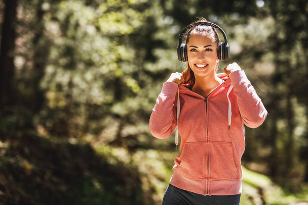 Young beautiful smiling female runner listening to music and taking a break after jogging in nature. - Photo, image