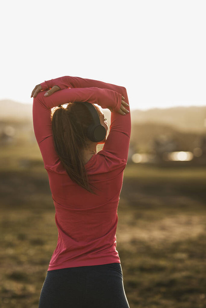 Young beautiful smiling female runner listening to music while stretching after jogging in nature. - Photo, Image