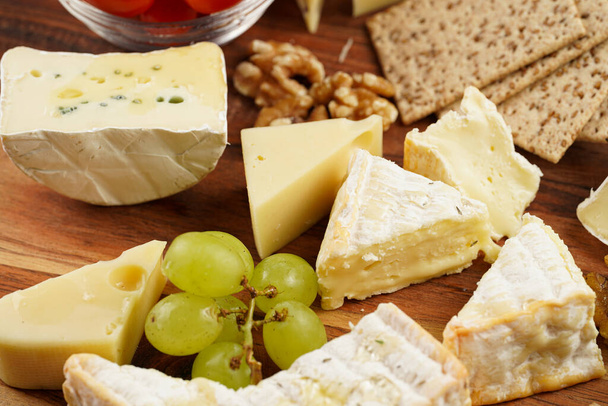 Πιάτο τυριού με βιολογικά τυριά - μπλε τυρί τσένταρ, emmantaler, γαλλικό μαλακό τυρί με έντονη μυρωδιά, ιταλική παρμεζάνα, σταφύλια, ντομάτες, ελιές, ξηρούς καρπούς και κράκερ σε ξύλινη σανίδα - Φωτογραφία, εικόνα