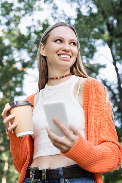 low angle view of happy woman holding paper cup and smartphone  - Foto, Imagem