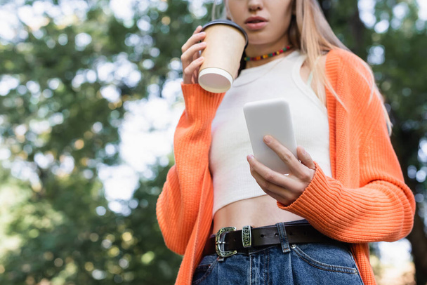 low angle view of cropped woman holding paper cup and smartphone in green park - Foto, immagini