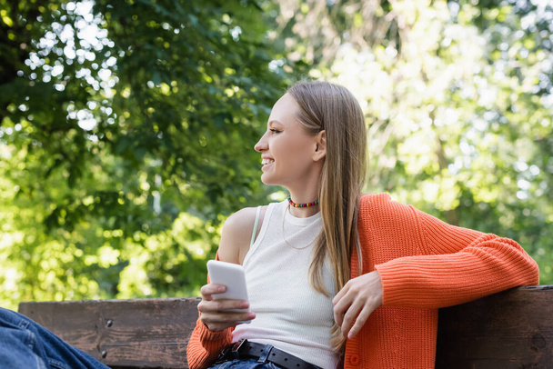 pleased woman woman holding smartphone and sitting on bench - Foto, imagen