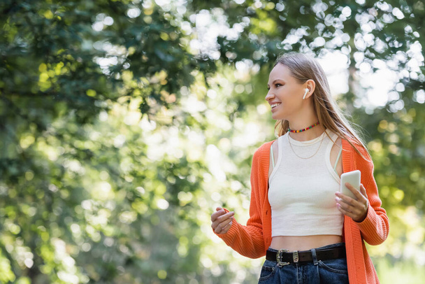 radostná žena v bezdrátovém sluchátku drží smartphone v parku - Fotografie, Obrázek