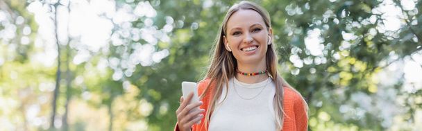 pleased woman in orange cardigan and wireless earphones using smartphone in park, banner - Foto, Imagen