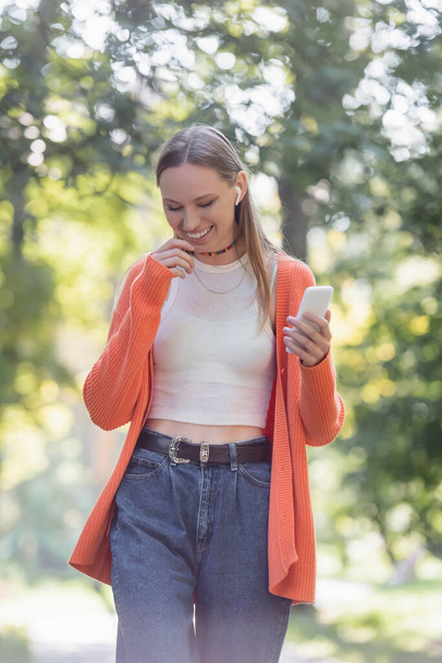 cheerful woman in orange cardigan and wireless earphone using smartphone in park - Foto, immagini