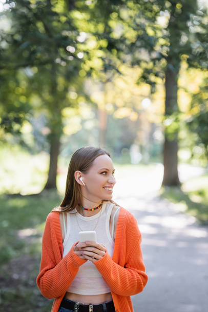 pleased woman in orange cardigan and wireless earphone using smartphone in park - Photo, image
