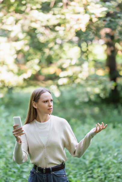 χαμένη νεαρή γυναίκα με το smartphone κοιτάζει μακριά, ενώ gesturing στο πάρκο  - Φωτογραφία, εικόνα