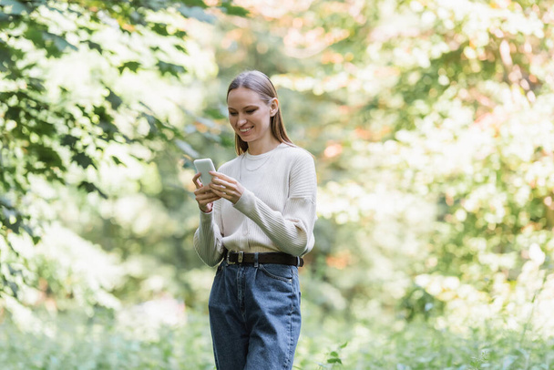 cheerful young woman with smartphone looking away in green park  - Foto, Imagen