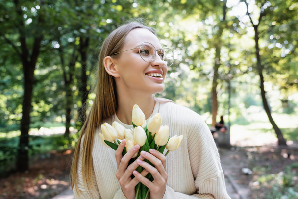 cheerful woman in glasses holding tulips in green park - Фото, зображення