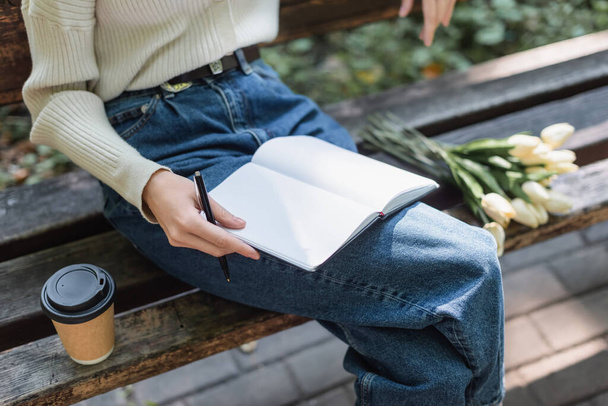 cropped view of woman in jeans holding notebook near bouquet of tulips and paper cup on bench - Foto, immagini