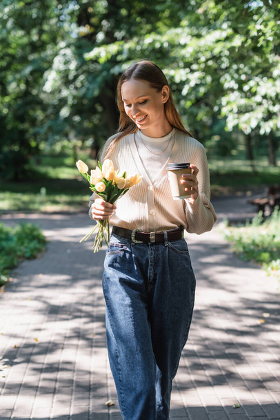 joyful woman in glasses and slouchy jeans walking with tulips and paper cup in green park - Photo, Image