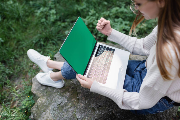 vysoký úhel pohledu šťastné ženy pomocí notebooku se zelenou obrazovkou při sezení na kameni  - Fotografie, Obrázek