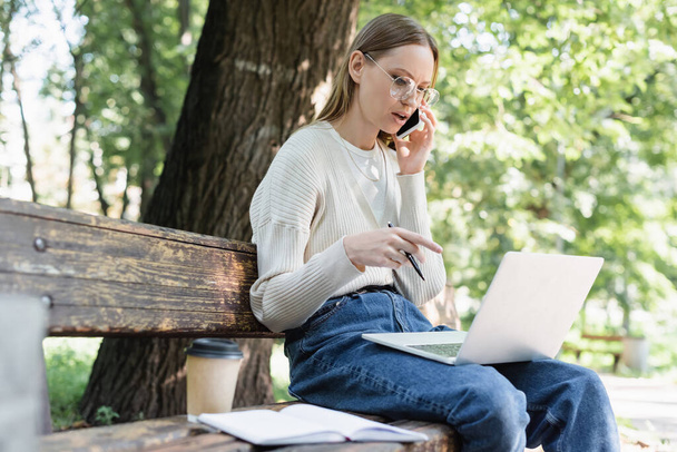 γυναίκα με γυαλιά μιλώντας στο smartphone και χρησιμοποιώντας φορητό υπολογιστή, ενώ κάθεται στον πάγκο στο πράσινο πάρκο - Φωτογραφία, εικόνα