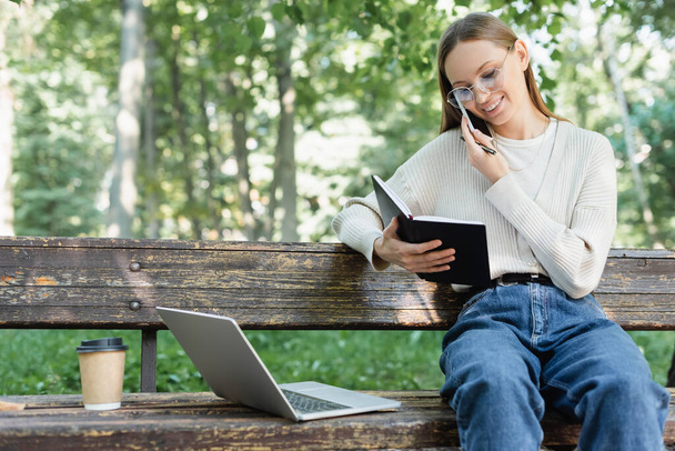 cheerful woman in glasses talking on smartphone while sitting near laptop on bench - Foto, Imagen