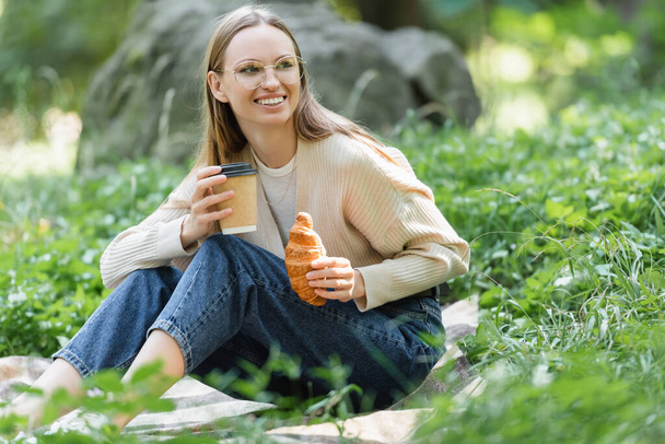 mujer sonriente en vasos sosteniendo taza de papel y croissant durante el picnic en el parque verde - Foto, imagen
