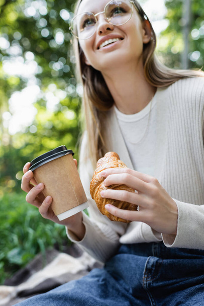 low angle view of happy woman in glasses holding paper cup and croissant in green park  - Photo, Image