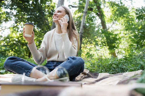 Gözlüklü mutlu kadın, kareli ekose üzerinde kağıt bardakla otururken akıllı telefonla konuşuyor. - Fotoğraf, Görsel
