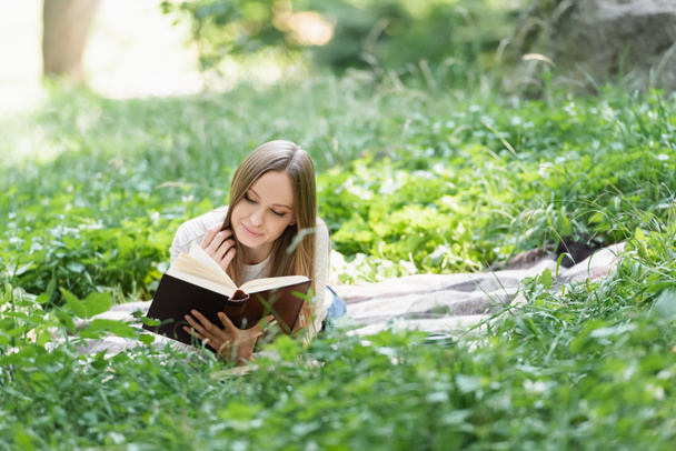 ευτυχισμένη γυναίκα ανάγνωση βιβλίο, ενώ βρίσκεται σε κουβέρτα γύρω από το πράσινο γρασίδι - Φωτογραφία, εικόνα