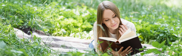 happy woman reading book while lying on blanket around green grass, banner - Фото, изображение