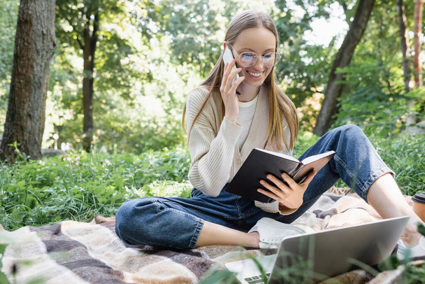 happy freelancer in glasses holding notebook while sitting on blanket and talking on smartphone near laptop  - Fotó, kép