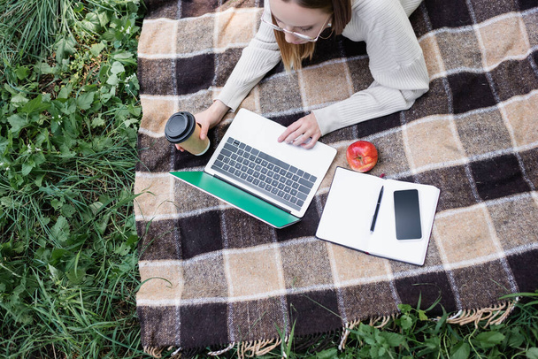 top view of freelancer in glasses lying on blanket and using laptop in park  - Fotó, kép