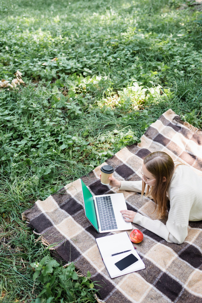 hoge hoek uitzicht op vrouw in bril liggend op deken en met behulp van laptop in park  - Foto, afbeelding