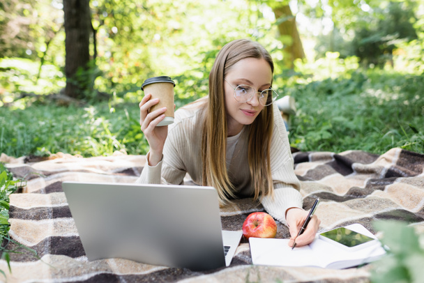 femme dans des lunettes prendre des notes tout en tenant tasse de papier près de gadgets sur couverture dans le parc - Photo, image