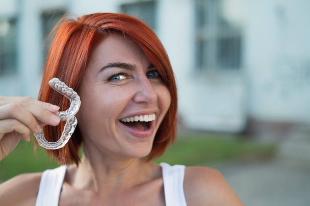 Red-haired Caucasian woman holding transparent mouthguards for bite correction outdoors. A girl with a beautiful snow-white smile uses silicone braces - Fotografie, Obrázek