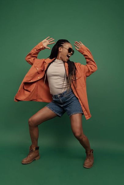 Excited african girl in shorts and shirt dancing, having fun isolated on dark green background. Concept of beauty, art, fashion, youth and emotions - Photo, image