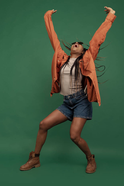 Emotional african girl in shorts and shirt dancing, having fun isolated on dark green background. Concept of beauty, art, fashion, youth and emotions - Fotoğraf, Görsel
