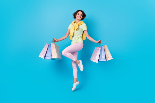 Full body photo of cute millennial brunette lady jump do shopping wear t-shirt pants sneakers isolated on blue background - Foto, Bild