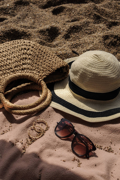 Αξεσουάρ παραλίας: ψάθινο καπέλο, καλοκαιρινή τσάντα, γυαλιά ηλίου και σκουλαρίκια στο κάλυμμα του κρεβατιού σε παραλία με άμμο μια καλοκαιρινή μέρα  - Φωτογραφία, εικόνα