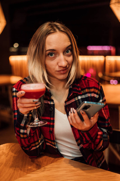 A young woman in a red shirt holds a glass of cocktail and a phone in her hand - Foto, Imagen