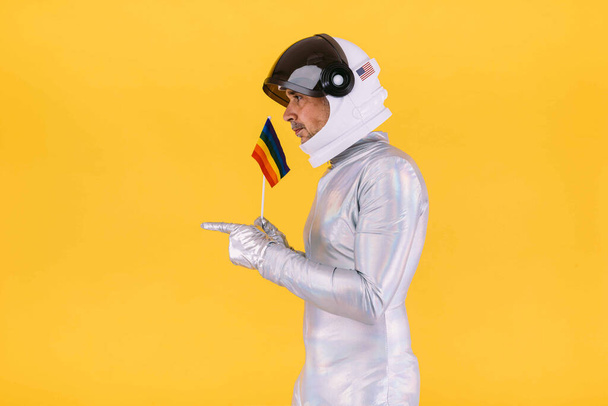 Гей чоловік одягнений як астронавт з шоломом і срібним костюмом, з прапором колективу lgtbi, вказуючи руками, на жовтому тлі. Гей, гомосексуалізм, транс, права і гендерна гордість концепція. - Фото, зображення