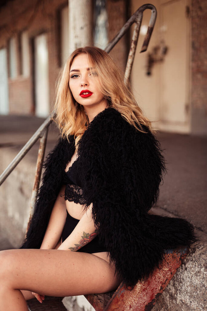 light blond girl with red lips in an eco-fur coat sits on the street and looks at the camera - Photo, Image