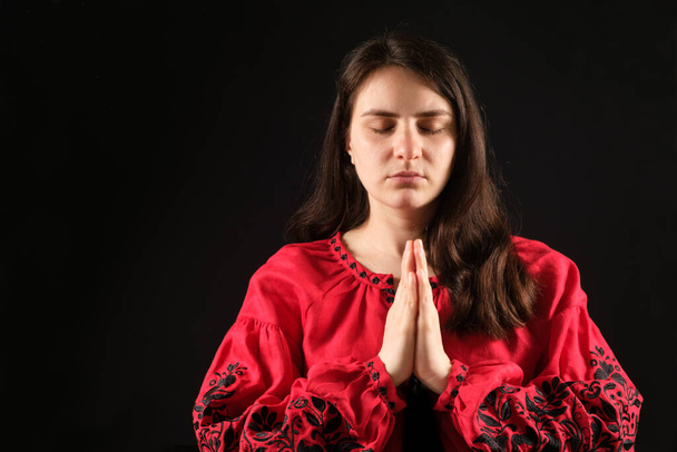 A young woman with her eyes closed in a red shirt folded her hands together in prayer, a black background, a place for text. - Photo, Image