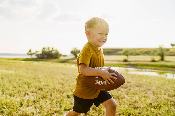 Blonde white boy smiling while playing with rugby ball outdoors - Foto, Bild