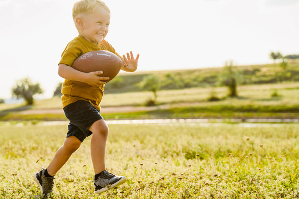 Blonde white boy smiling while playing with rugby ball outdoors - Фото, изображение