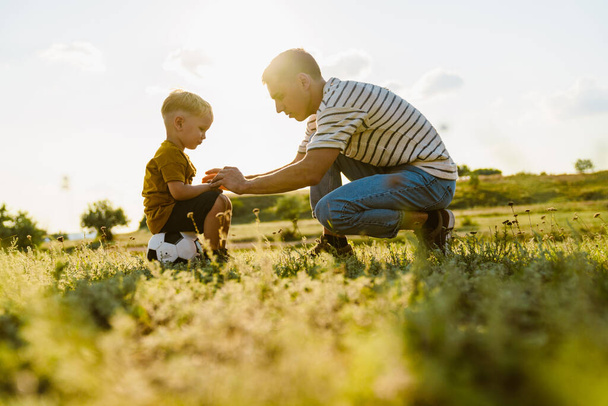 Joven padre blanco hablando con su triste hijo mientras se agacha en el campo de verano - Foto, imagen