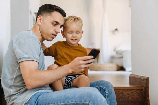 Young father and son using mobile phone while sitting on stairs at home - Foto, Imagem