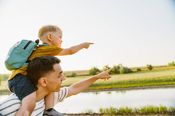 Young father gesturing while carrying little son on his shoulders outdoors - Photo, image