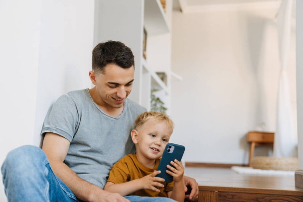 Young father and son smiling and using mobile phone at home - Φωτογραφία, εικόνα