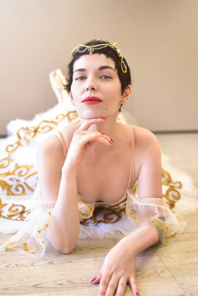 A portrait of a charming ballerina lying on the floor in a ballroom, she touches her chin with her hand and looks flirtatiously at the camera - 写真・画像