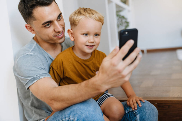 Young father and son smiling and taking selfie photo on mobile phone at home - Fotografie, Obrázek