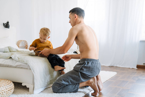 Mladý otec bez košile čte knihu, zatímco obléká svého syna doma - Fotografie, Obrázek