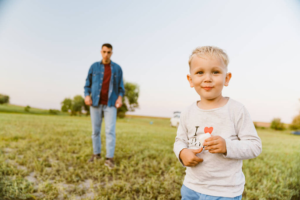 Blonde boy looking at camera while standing with her father on summer field - Fotoğraf, Görsel