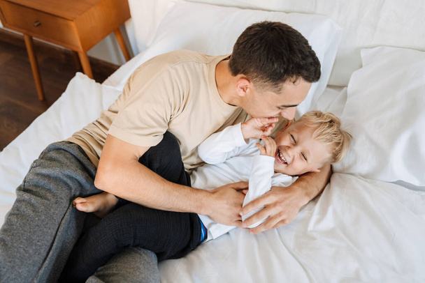 Young white father making fun and playing with his son on bed at home - Фото, изображение