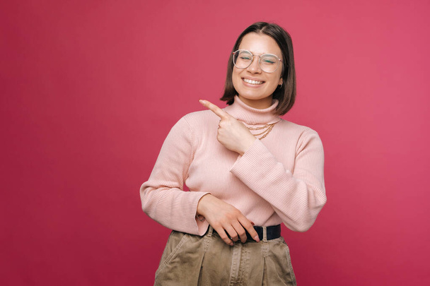 Young cheerful woman is pointing aside with a finger to the copy space. Studio shot over pink background. - Фото, изображение