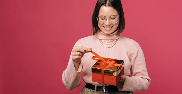 Panorama photo of a young woman is holding a red wrapped gift box. Studio shot over pink background. - 写真・画像